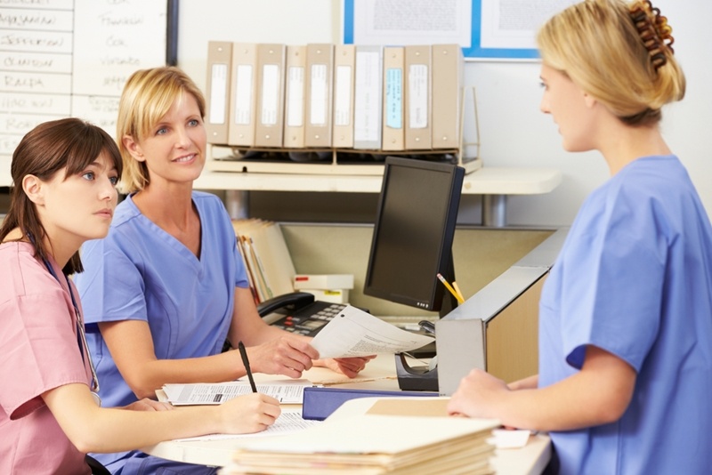 Three nurses dressed in scrubs sitting at nurses' station with piles of folders and papers. 